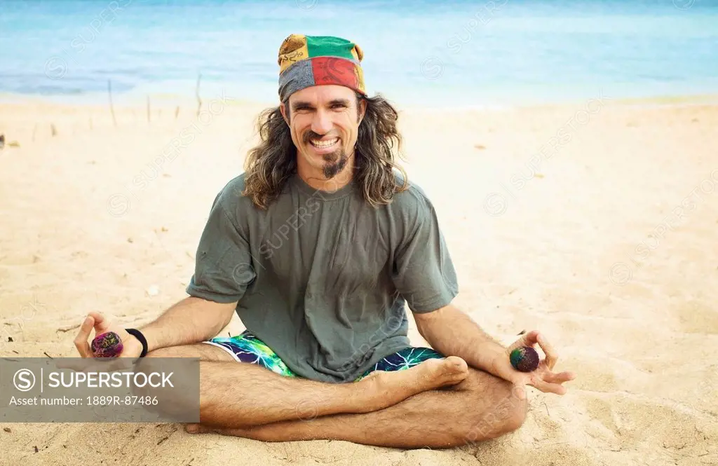 A Man Sits In The Sand In A Yoga Pose Holding Two Hackey Sacks;Hawaii United States Of America