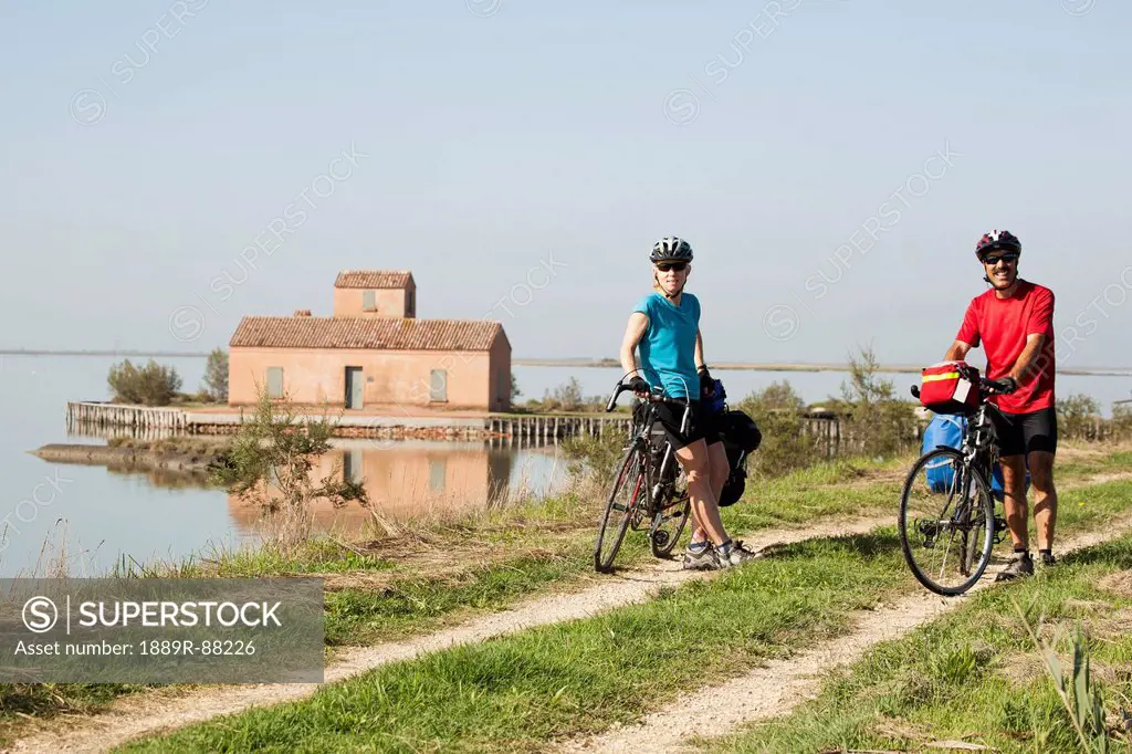 A Couple With Touring Bikes Along A Path And Body Of Water With A Boathouse In The Background;Comacchio Emilia-Romagna Italy