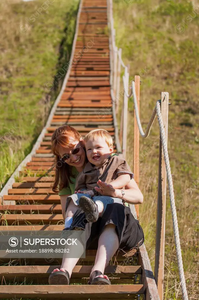 A mother and her young son sit together laughing on the lower steps of a long stairway leading to the top of a ski-jump at hilltop ski area;Anchorage ...