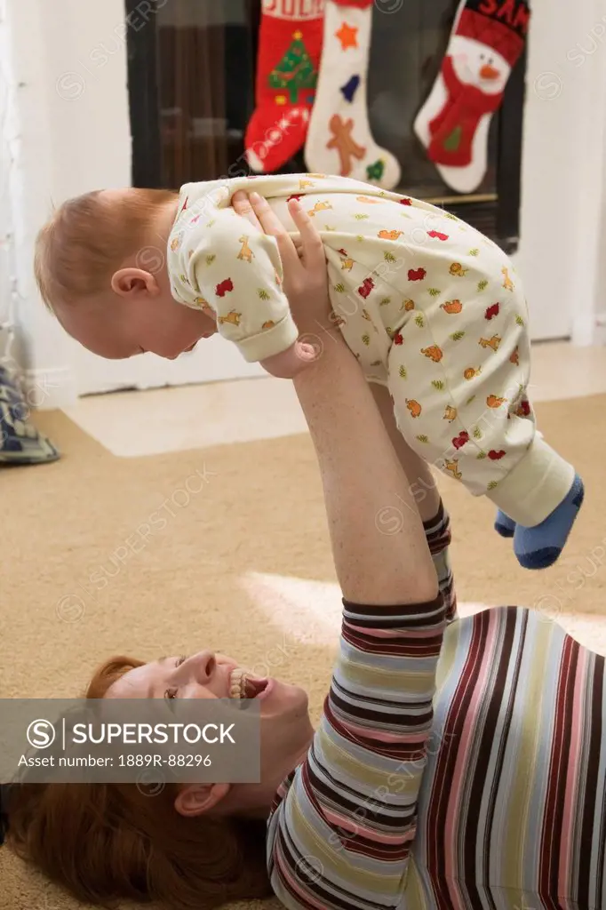 A mother laying on the floor and playing with her infant son with christmas stockings hung over the fireplace in the background;San diego california u...