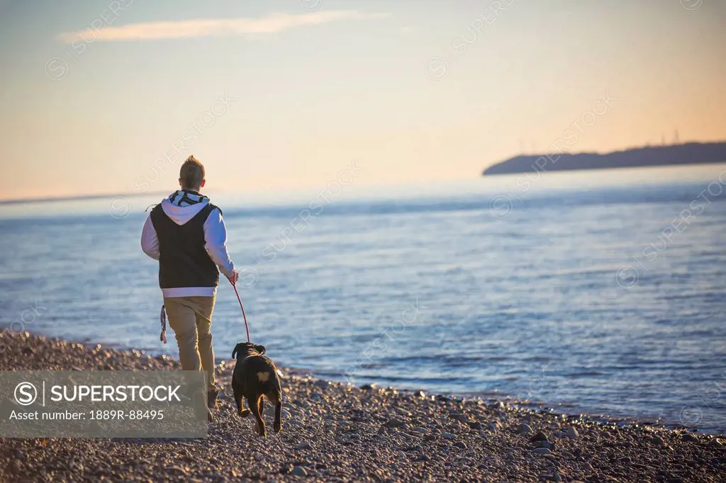 Teenage boy running with his rottweiler dog on the beach at point wornzof cook inlet and fire island in the background at sunset;Anchorage alaska unit...