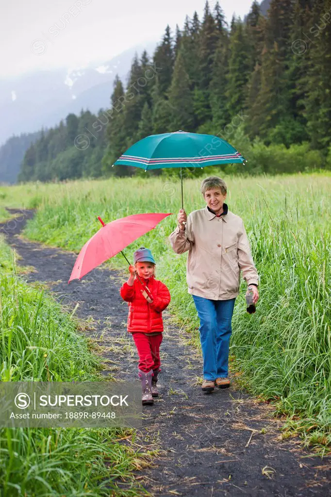 Grandmother and granddaughter playing with their umbrella while walking up the path from the beach at Lowell Point on a rainy day, summer, Seward, Sou...