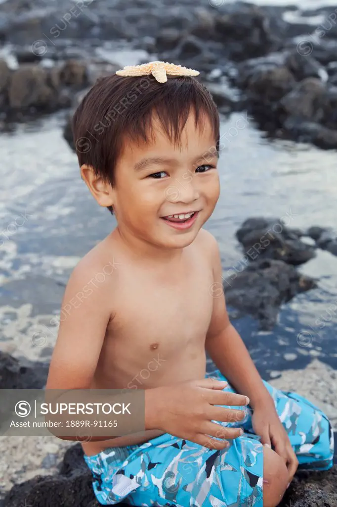 A young boy sits at the water's edge with a starfish on his head;Honolulu oahu hawaii united states of america