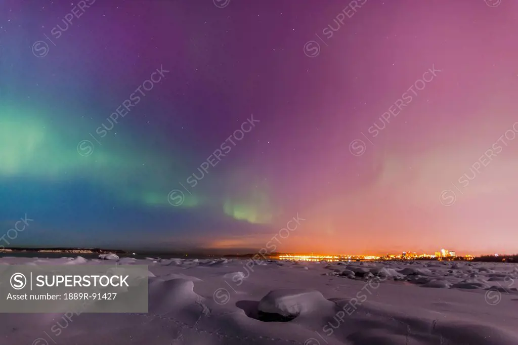The Northern Lights shine above the Anchorage city skyline in this nighttime view from the Tony Knowles Coastal Trail in winter; Anchorage, Alaska, Un...