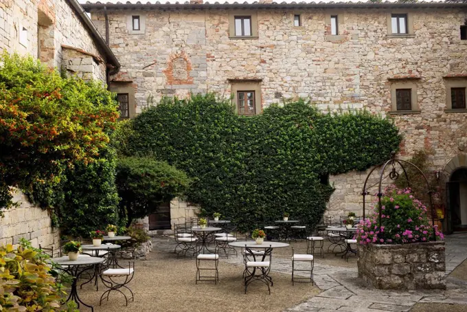 Courtyard filled with tables and chairs, vines and shrubs growing around stone buildings; Gaiole in Chianti, Toscana, Italy