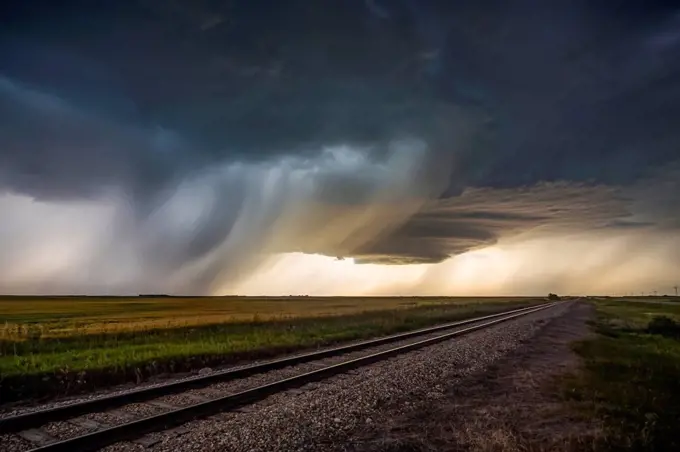 Dark storm clouds over railroad tracks in a field with rain falling in the distance; Marquis, Saskatchewan, Canada