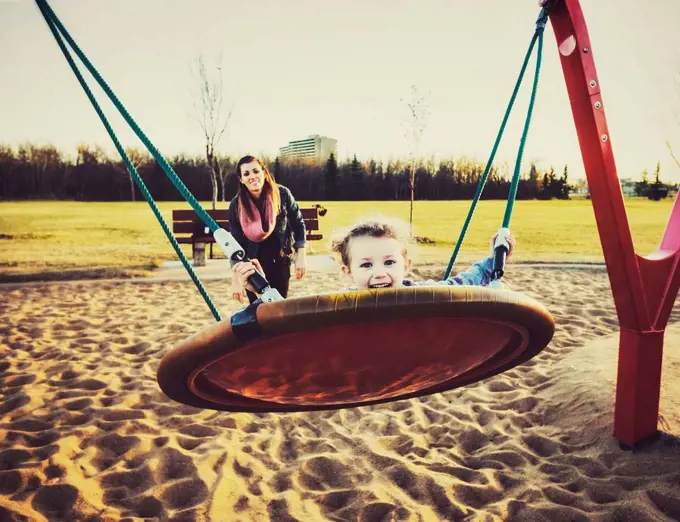 A young mom and her daughter playing on a saucer swing in a playground on a warm autumn evening; Edmonton, Alberta, Canada