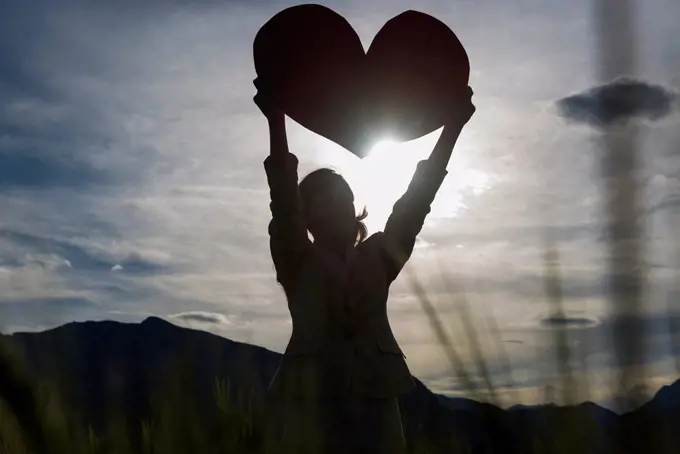 Silhouette of a woman holding up a large heart shape against the sunlight