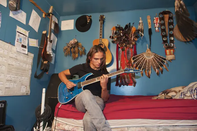 Indigenous young man playing guitar in bedroom; Rossburn, Manitoba, Canada