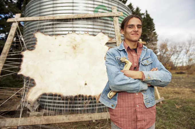 Indigenous young man standing in front of a stretched Bison hide; Rossburn, Manitoba, Canada