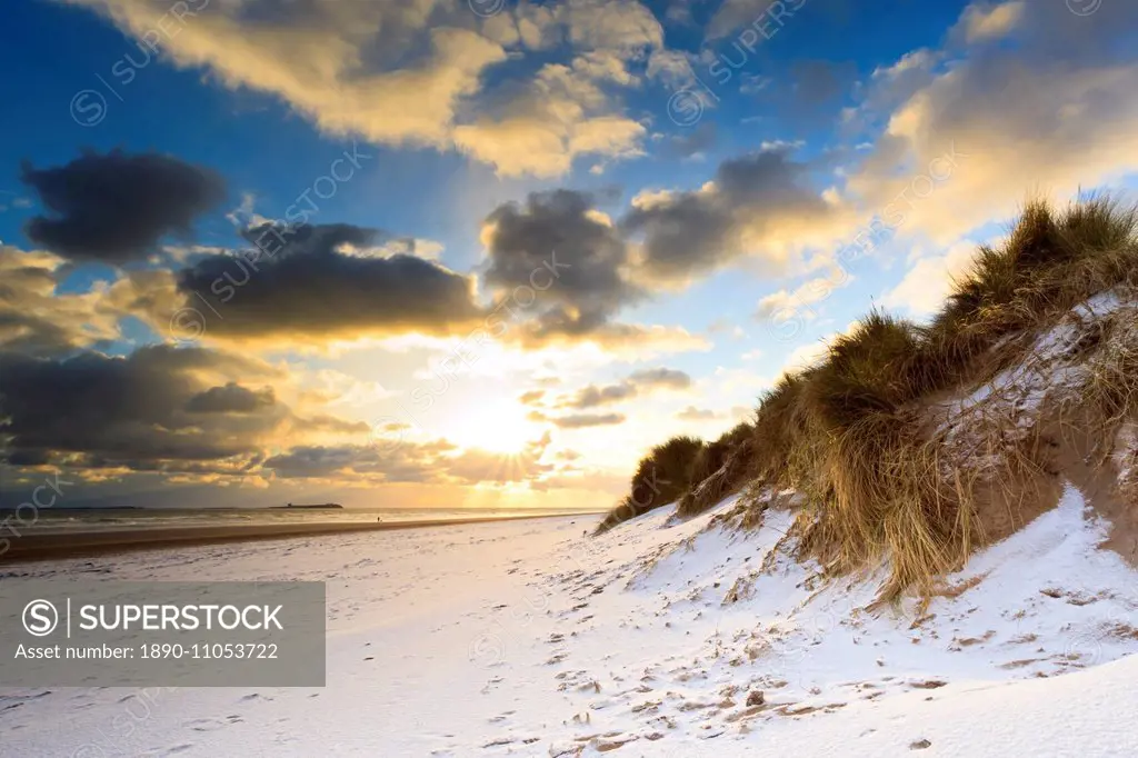 Man walks dog on snow covered Bamburgh beach at dawn with view to Farne Islands, Northumberland, England