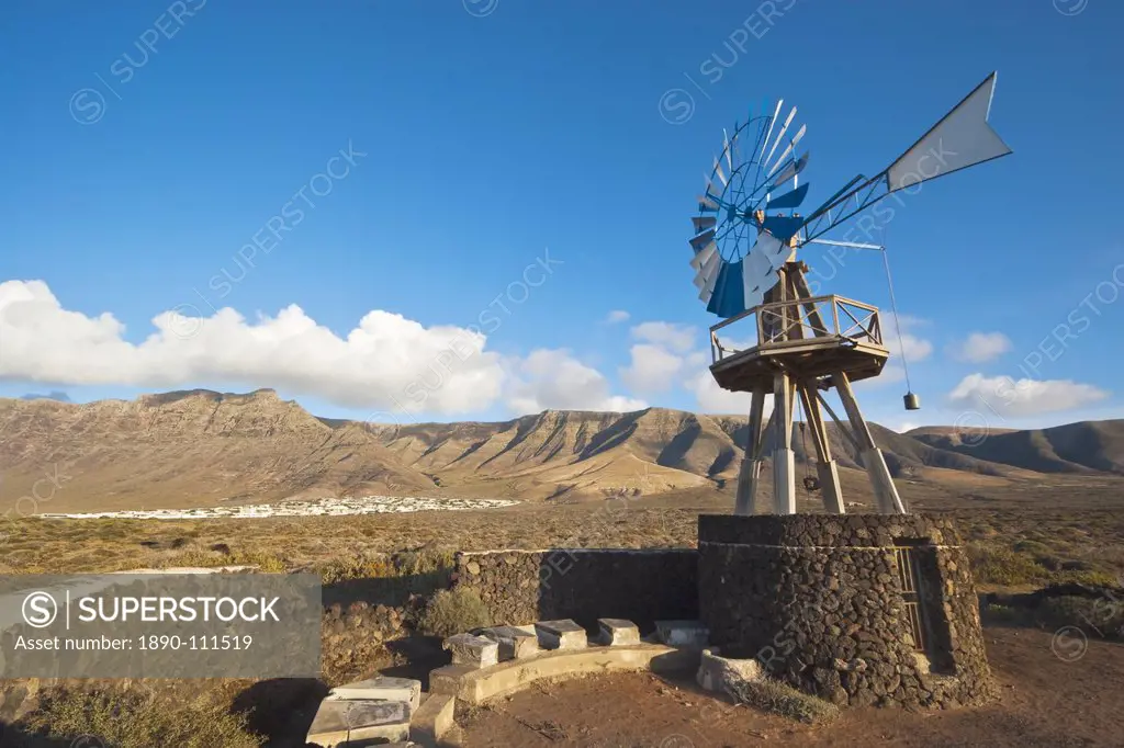 Traditional wind pump with 600m volcanic cliffs of the Risco de Famara rising over desert landscape near Famara in the north west of the island, Famar...