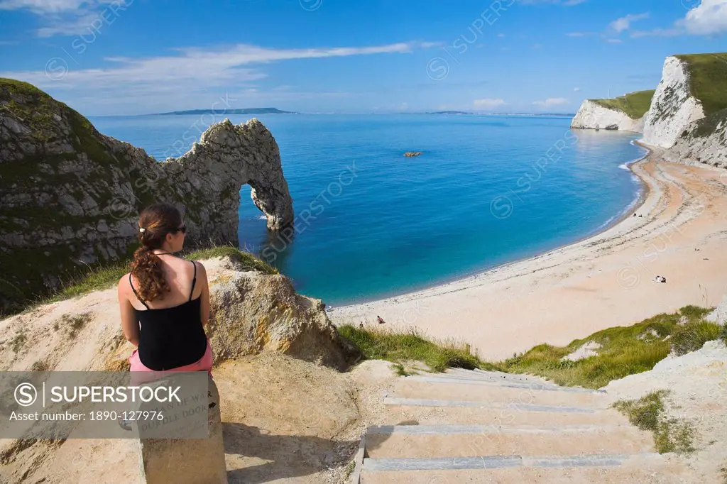 Woman resting on a clifftop sign, looking towards Durdle Door beach, Jurassic Coast, UNESCO World Heritage Site, Dorset, England, United Kingdom, Euro...