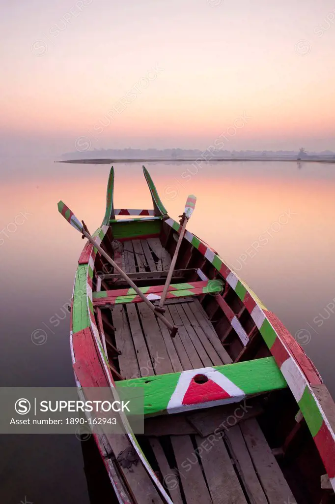 Traditional rowing boat moored on the edge of flat calm Taungthaman Lake at dawn with the colours of the sky reflecting in the calm water, close to th...