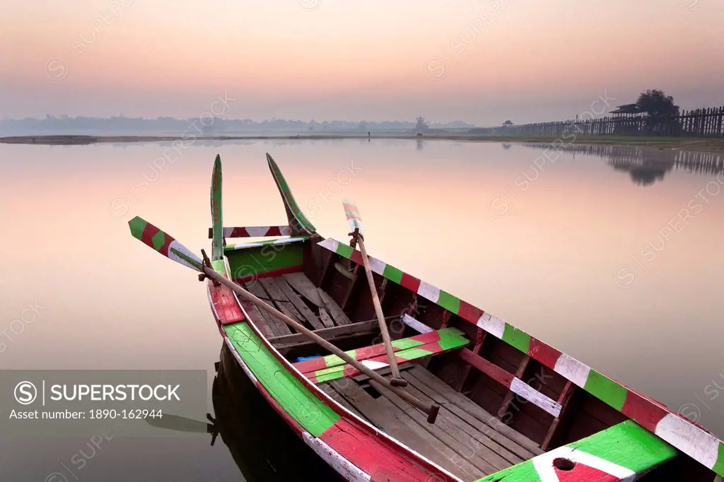 Traditional rowing boat moored on the edge of flat calm Taungthaman Lake at dawn with the colours of the sky reflecting in the calm water, close to th...