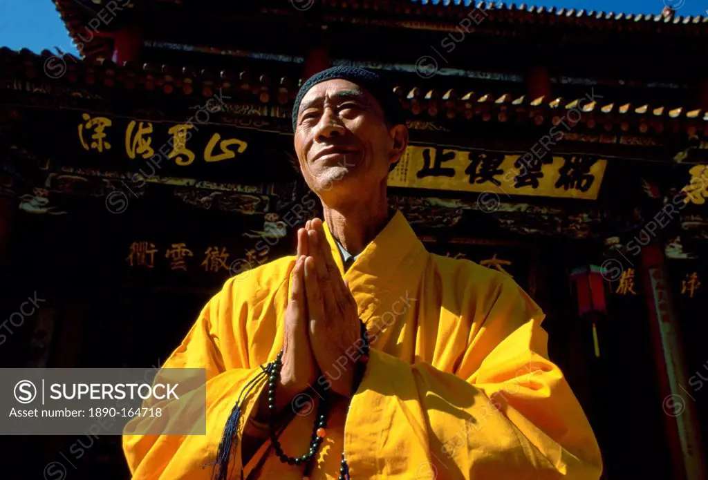 A Buddhist monk in saffron coloured robes with hands together while praying at the Buddhist Temple in Huating, China