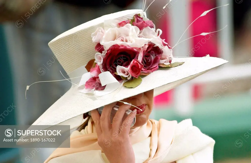 Race-goer wearing a cream hat trimmed with flowers at Royal Ascot Races while chatting on her mobile cell phone.