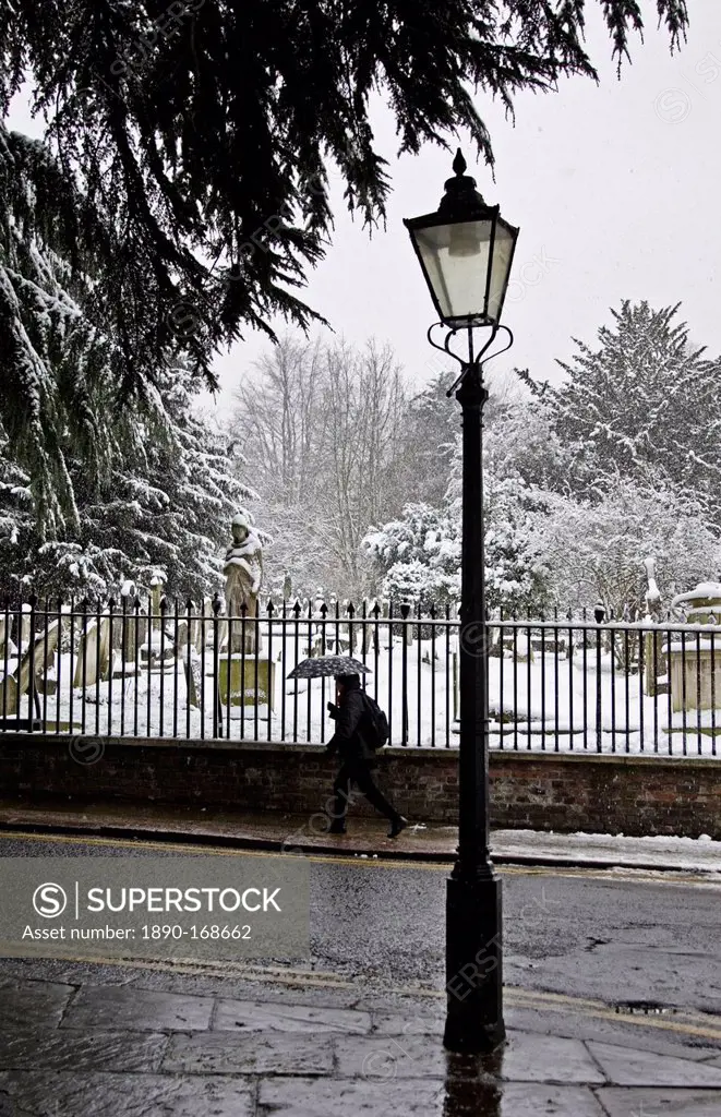 Woman with umbrella walks past snow covered graveyard, Hampstead, North London, United Kingdom