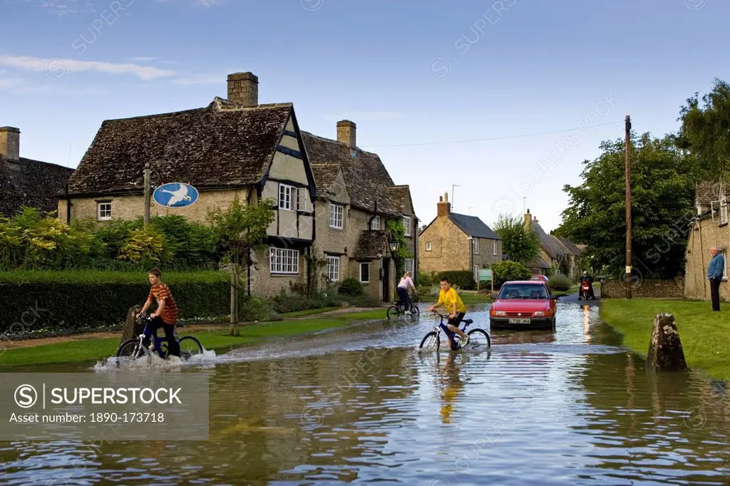 Children ride their bikes through flood water in Minster Lovell, The Cotswolds, Oxfordshire, England, United Kingdom