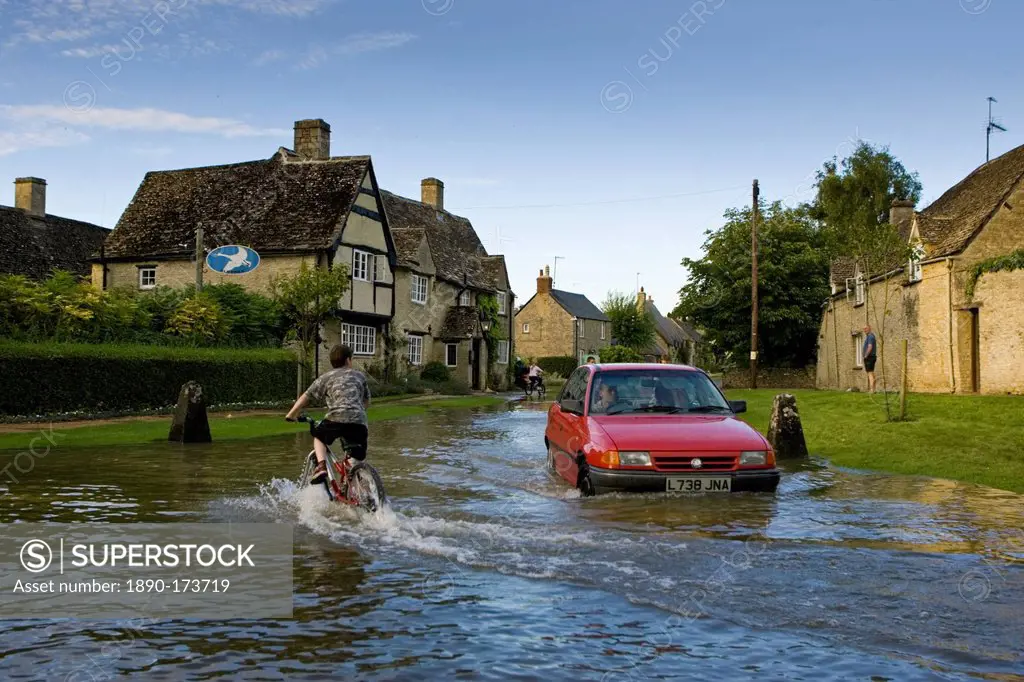 Car and boy on a bike try to move through flood water in Minster Lovell, Oxfordshire, England, United Kingdom
