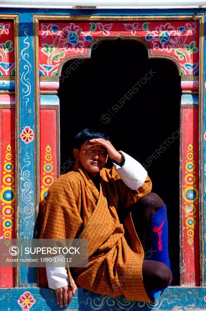 Bhutanese man shielding his eyes from the sun as he sits in an ornate carved and decorated window. His modern Nike sports socks contrast with his trad...