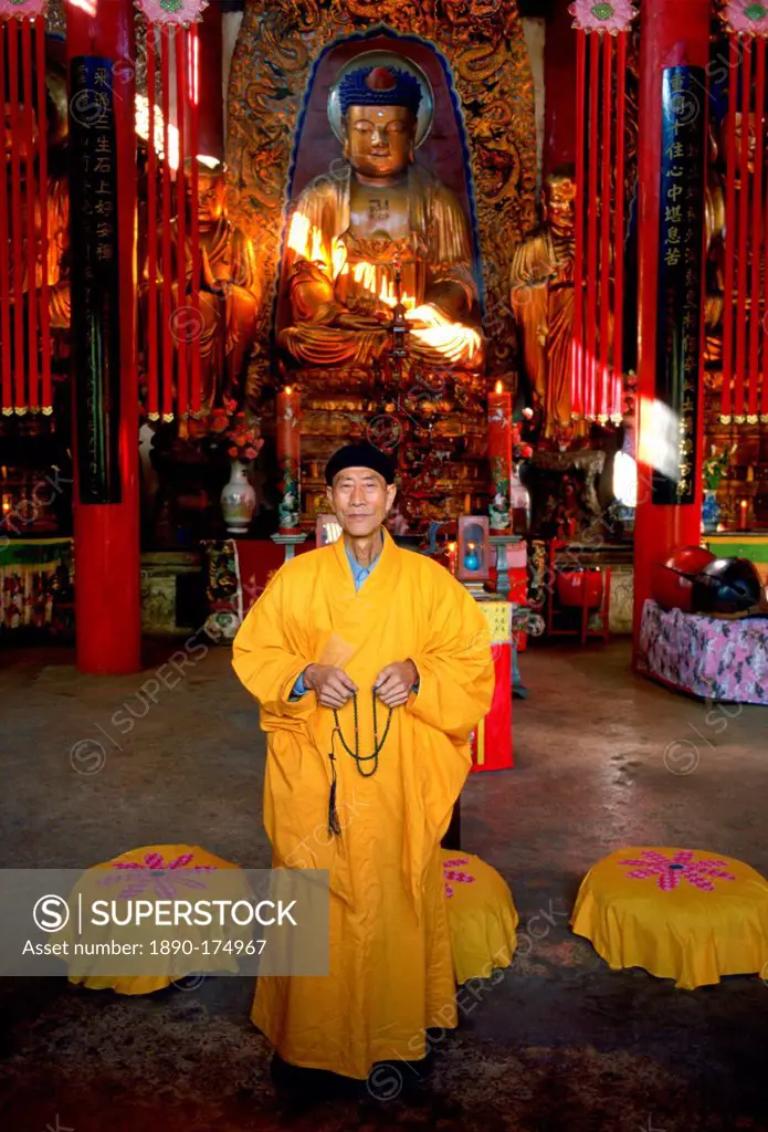 A Buddhist monk in saffron coloured robes praying in front of a statue of Buddha at the Buddhist Temple in Huating, China