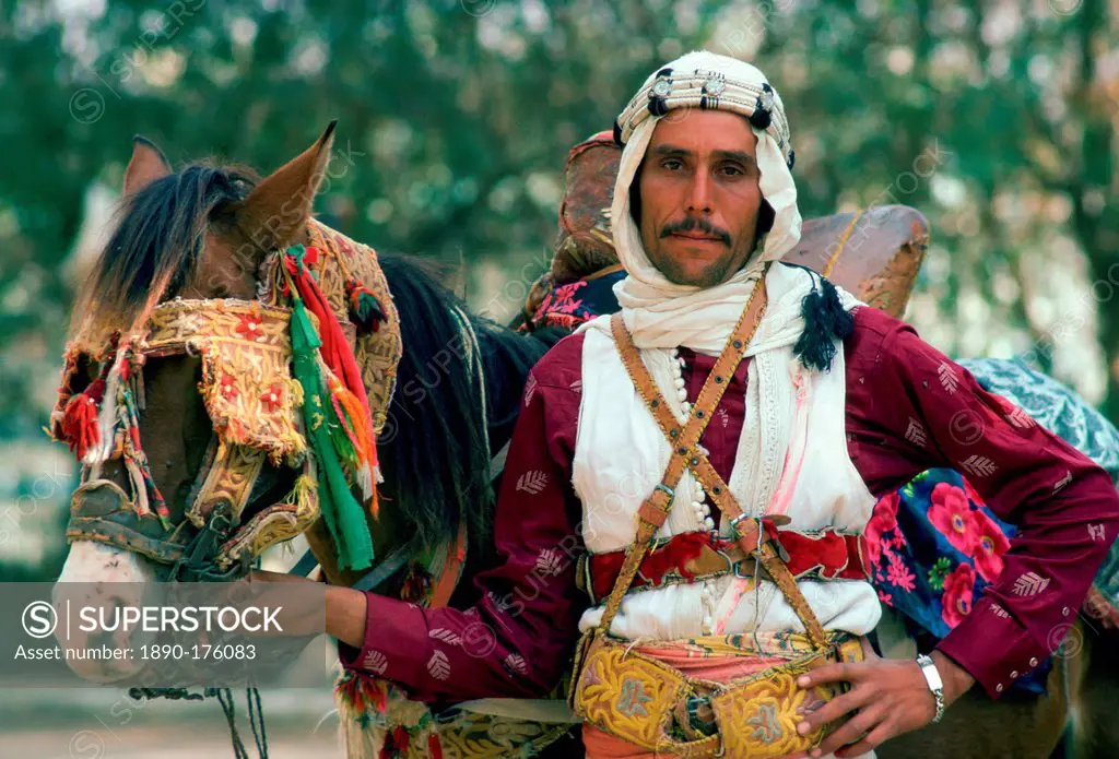 A horseman in traditional dress holding his horse which has eye defenders against the sun and dust.