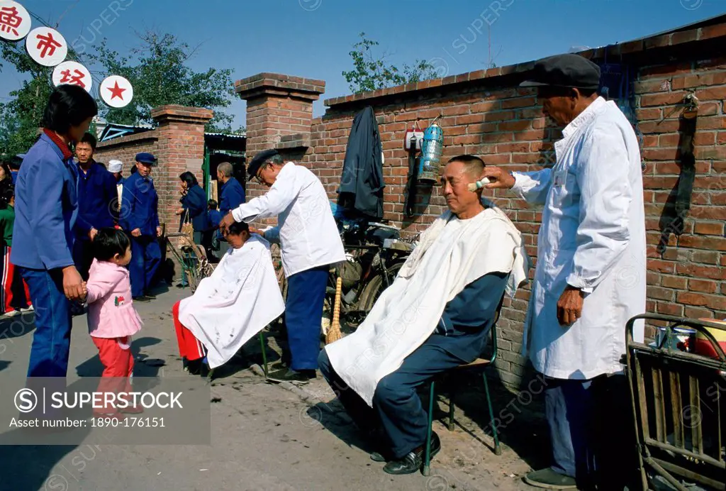 Men at work at an outdoor Barbers' Shop in Old Peking (Beijing), China. One is using a shaving brush with foam and the other is cutting a young boy's ...