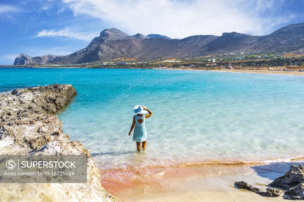 Woman in dress standing in the turquoise clear sea facing the pink sand beach of Falassarna, Crete, Greek Islands, Greece, Europe