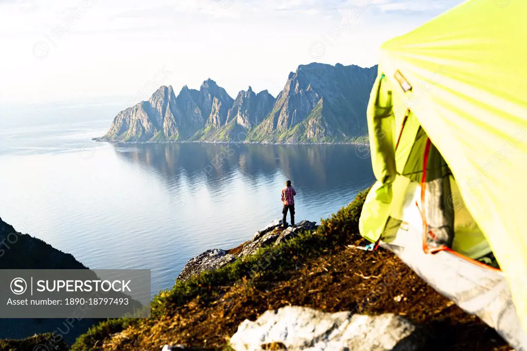 Hiker admiring mountains standing on top of cliff beyond camping tent, Senja island, Troms county, Norway, Scandinavia, Europe