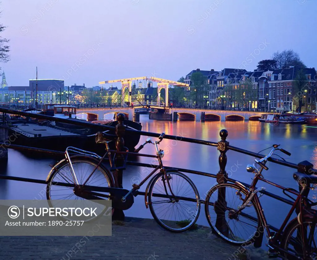 Bicycles by the side of the canal, with barges, and the Magere Brug Skinny Bridge in the background, in the evening in Amsterdam, Holland, Europe