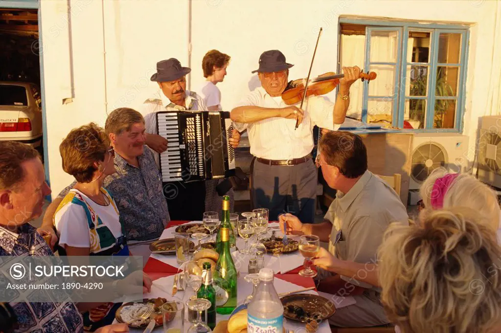 Tourists in open air restaurant being entertained by musicians including man playing accordion on Mykonos, Cyclades Islands, Greek Islands, Greece, Eu...