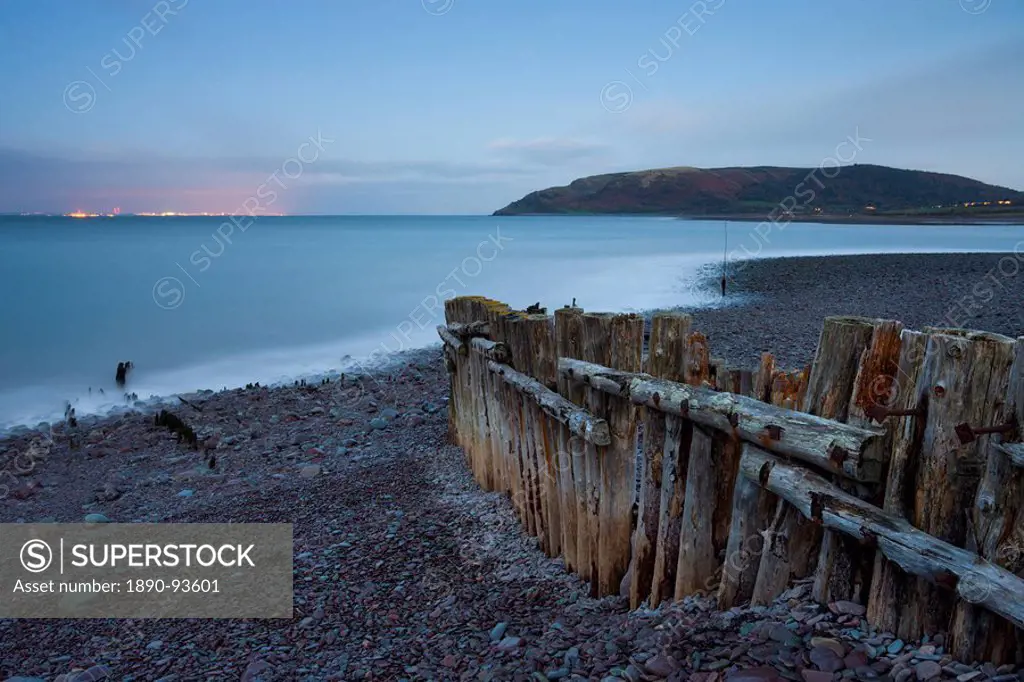 Weathered coastal defences at Porlock Weir in Exmoor National Park, with the evening lights of Wales visible across the Bristol Channel, Somerset, Eng...