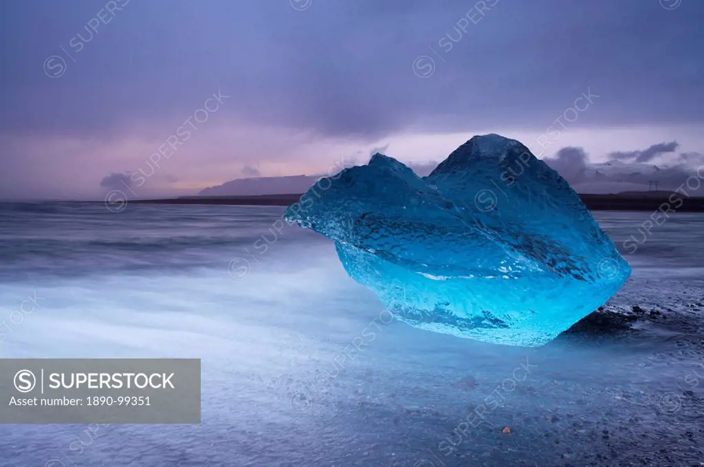 Translucent blue iceberg washed ashore on Breidamerkursandur black sands, near Jokulsarlon glacial lagoon, Oraefajokull Vatnajokull glacier in the dis...