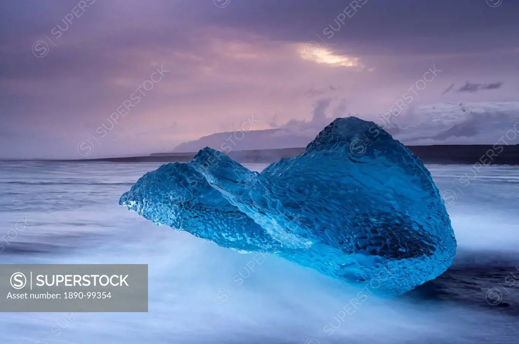 Translucent blue iceberg washed ashore on Breidamerkursandur black sands, near Jokulsarlon glacial lagoon, Oraefajokull Vatnajokull glacier in the dis...