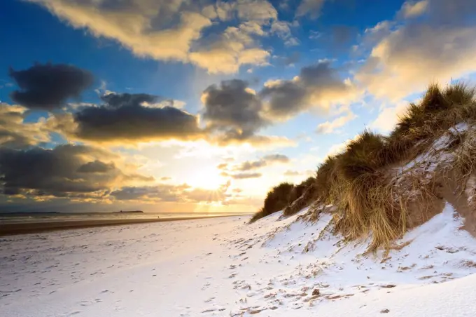 Man walks dog on snow covered Bamburgh beach at dawn with view to Farne Islands, Northumberland, England