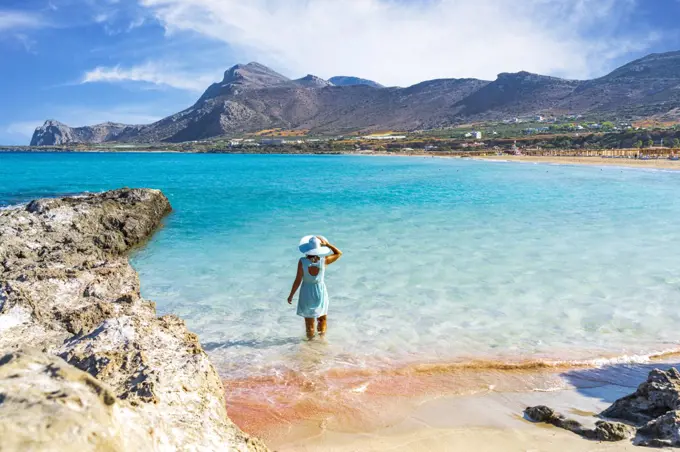 Woman in dress standing in the turquoise clear sea facing the pink sand beach of Falassarna, Crete, Greek Islands, Greece, Europe