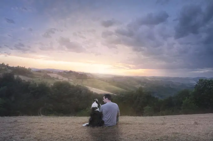 A man and his border collie dog sitting in a field in the countryside enjoying sunset, Italy, Europe