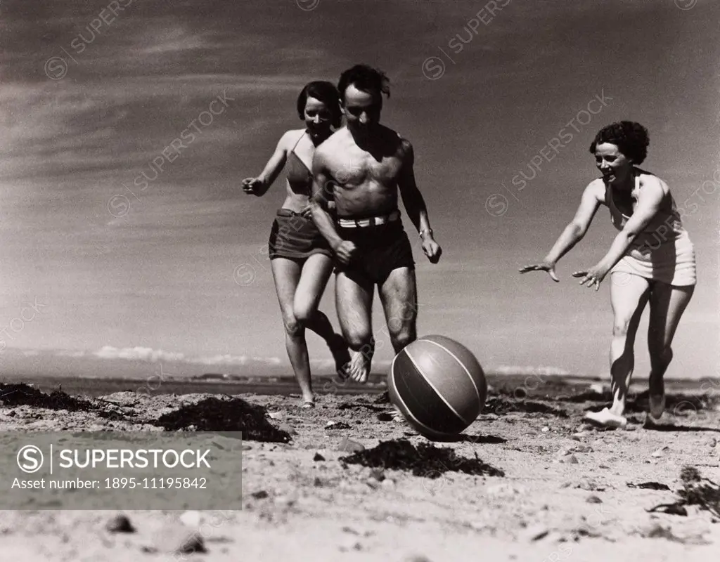 Two women and a man chasing a beach ball along the beach, c 1930s.