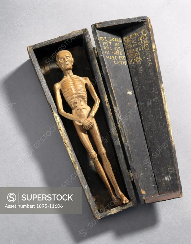A carved wooden statue of a dead man in a miniature coffin. The figure is emaciated and almost skeletal. There is a latin inscription in the lid of th...