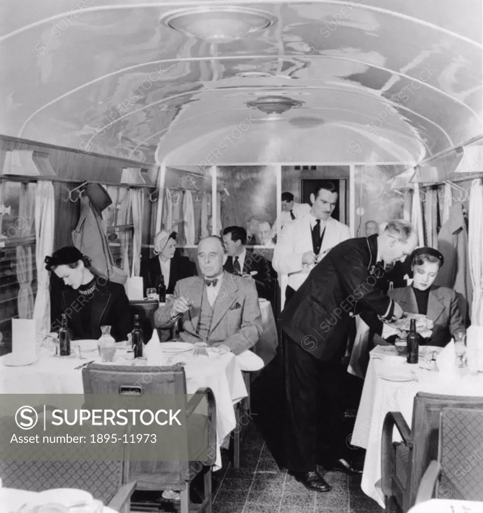 Stewards in British Railways First Class dining car procede down the car with platters of food from which they serve passengers. Posed with models.