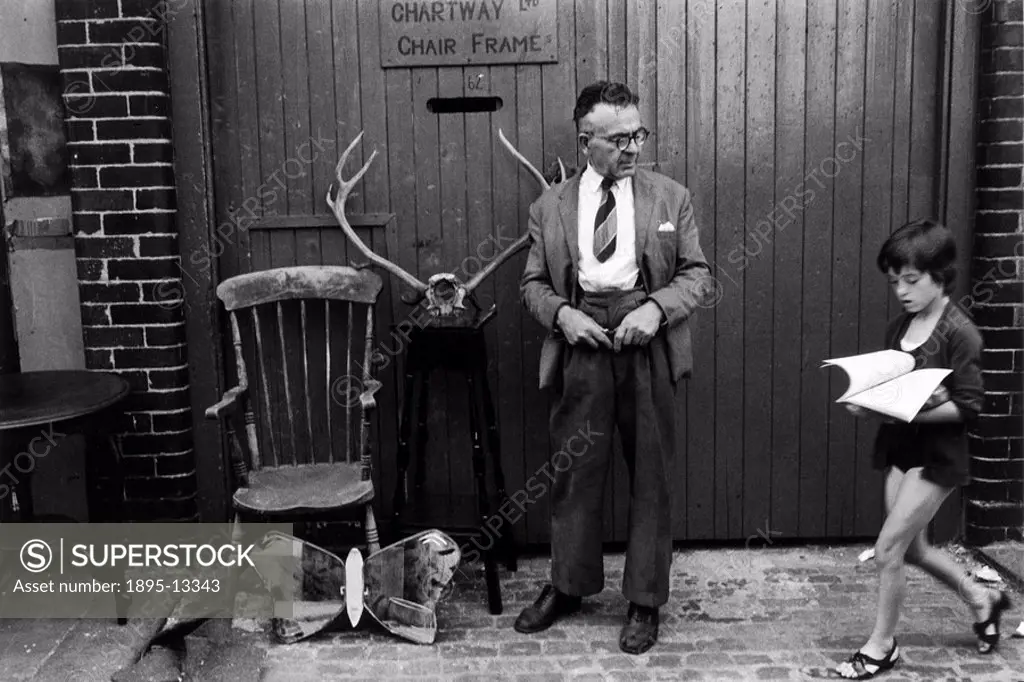 Man with antique bric_a_brac, Wormwood Scrubs, London, 1967. A man standing with antique bric_a_brac outside a lock_up garage, watching a young girl w...