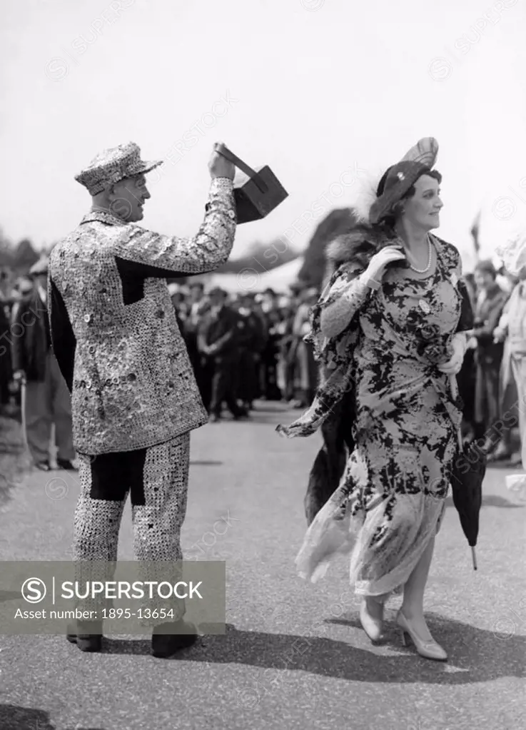A traditional London Pearly King in his suit covered in mother-of-pearl buttons collects money in the enclosure at Ascot Races. Walking past is a woma...
