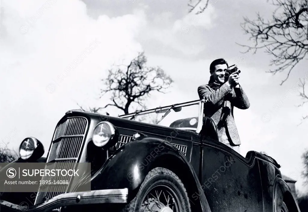 Man standing in a Ford motor car, filming with a cine camera, c 1930s.Innovations in the design of home movie cameras meant that the cameras were smal...