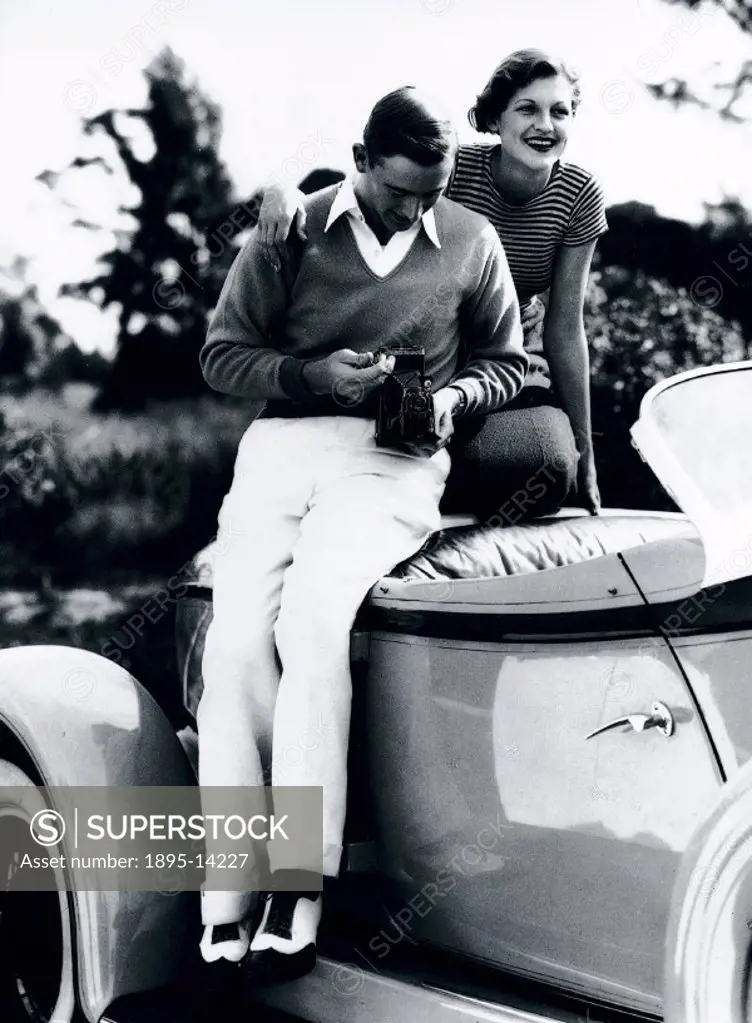 Young man holding a Kodak Series III camera, sitting on an American sports coupe with his girlfriend.