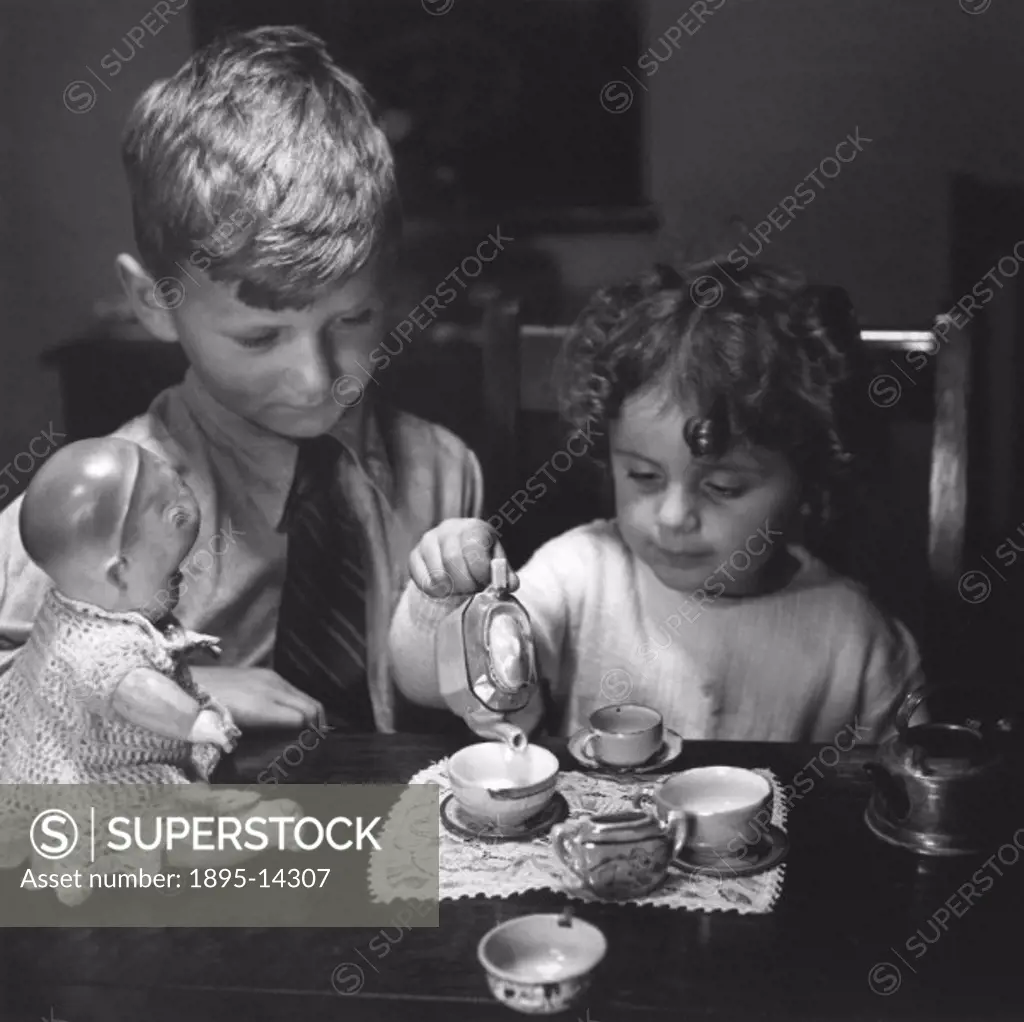 Young girl pouring tea as her brother looks on, c 1930s.