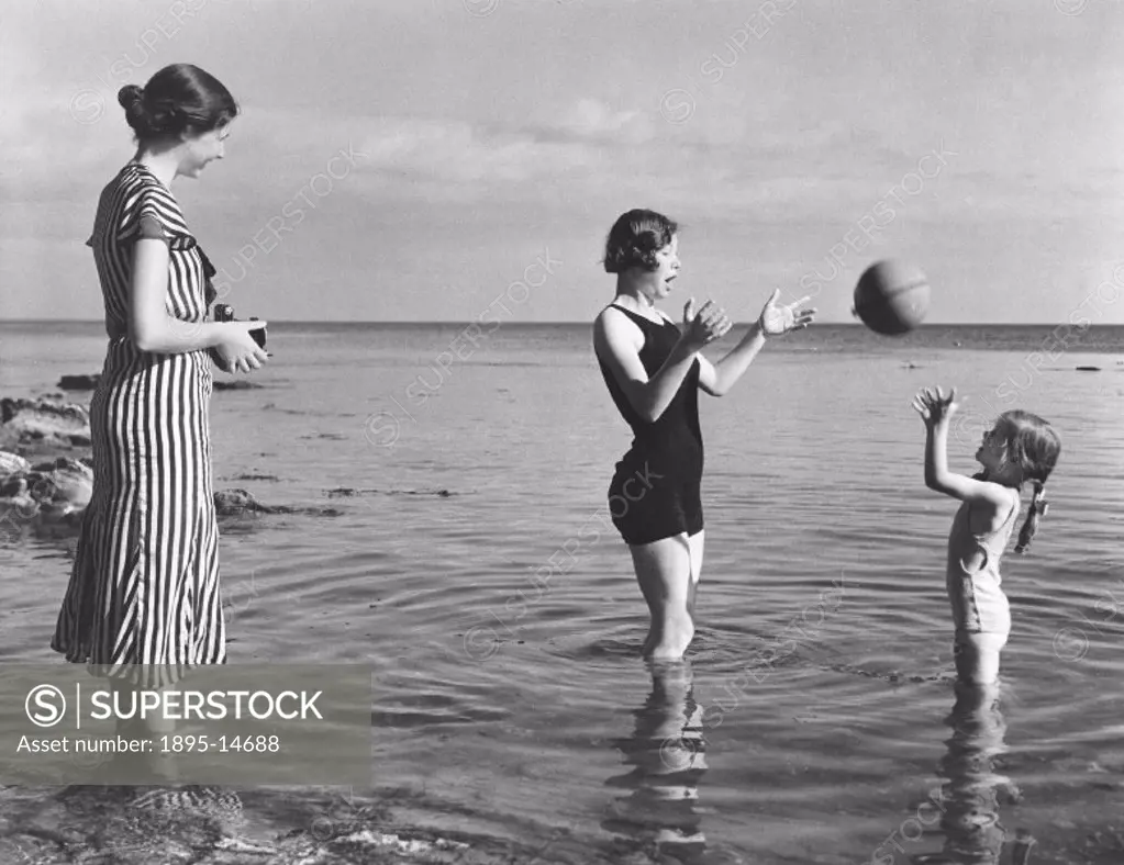 Woman wearing the latest fashions taking a photograph of a mother and child playing with a ball in the sea.
