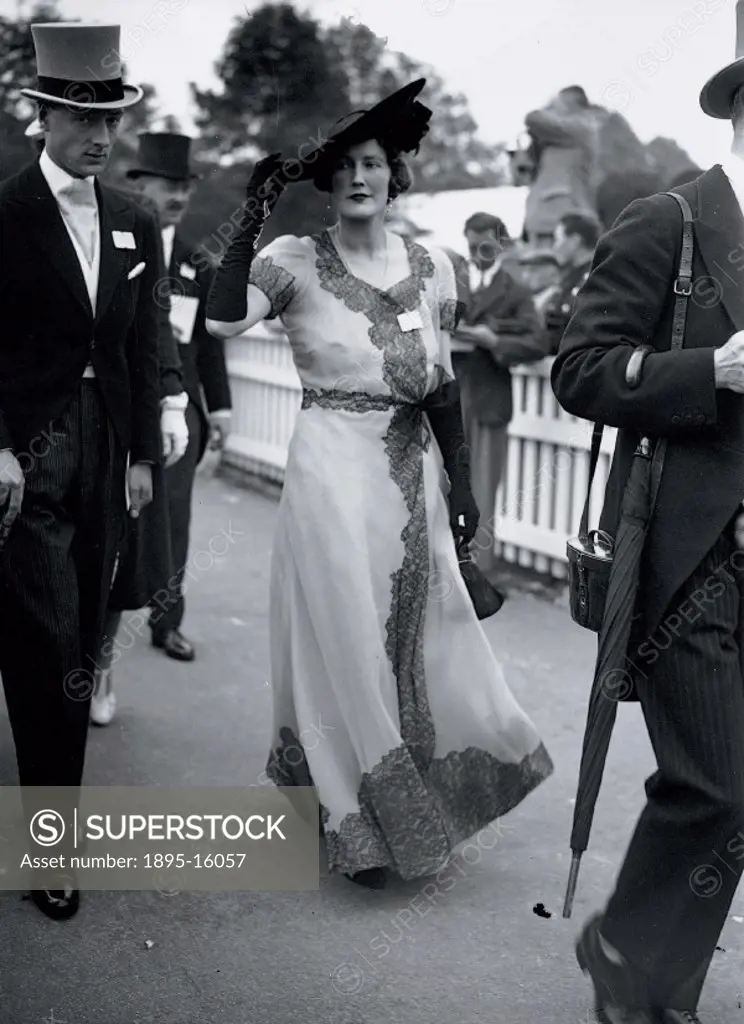 A couple walking through the enclosure on day two of the Ascot races in the fashions of the summer. She is wearing a long flared cross-over dress dec...
