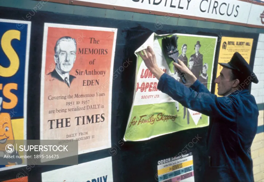 A billposter is shown pasting posters on the walls of a platform in Piccadilly Circus station, including one advertising the serialisation of the memo...