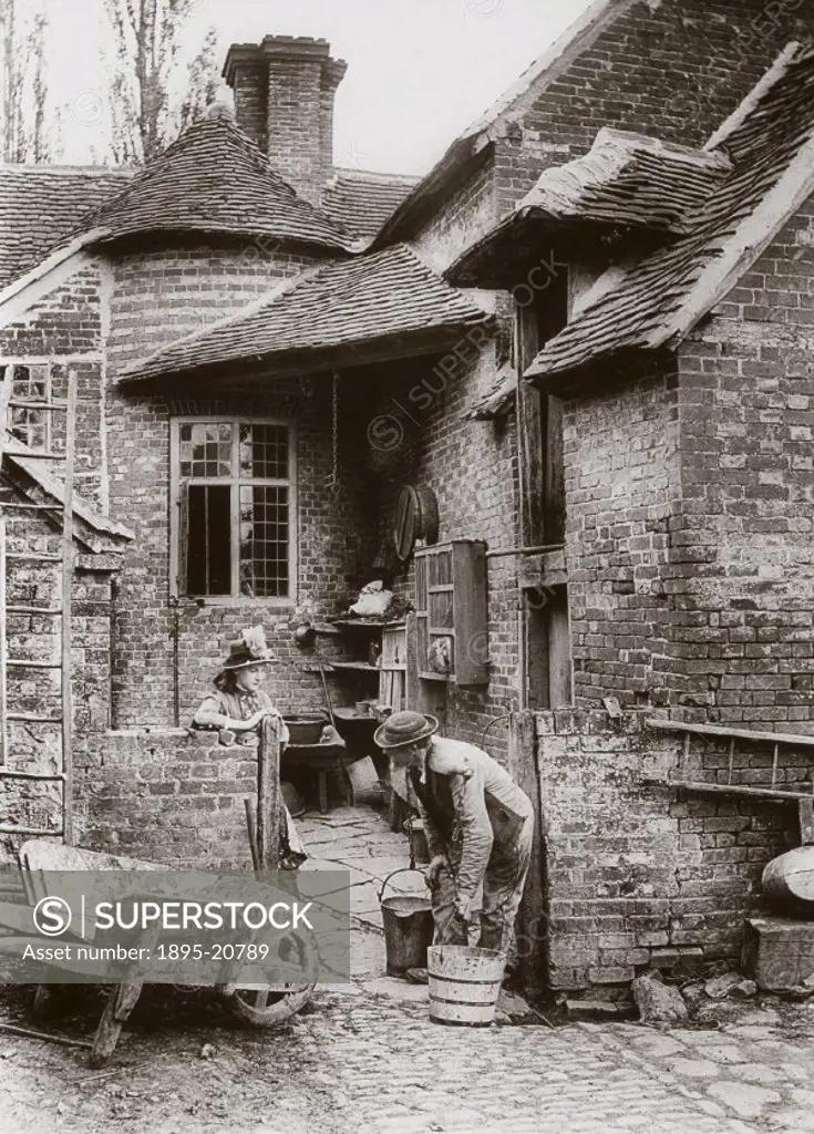 Photograph by Colonel Gale, showing a man with a yoke on his shoulders about to lift two milk pails. A woman in a hat is standing beside the yard gate...